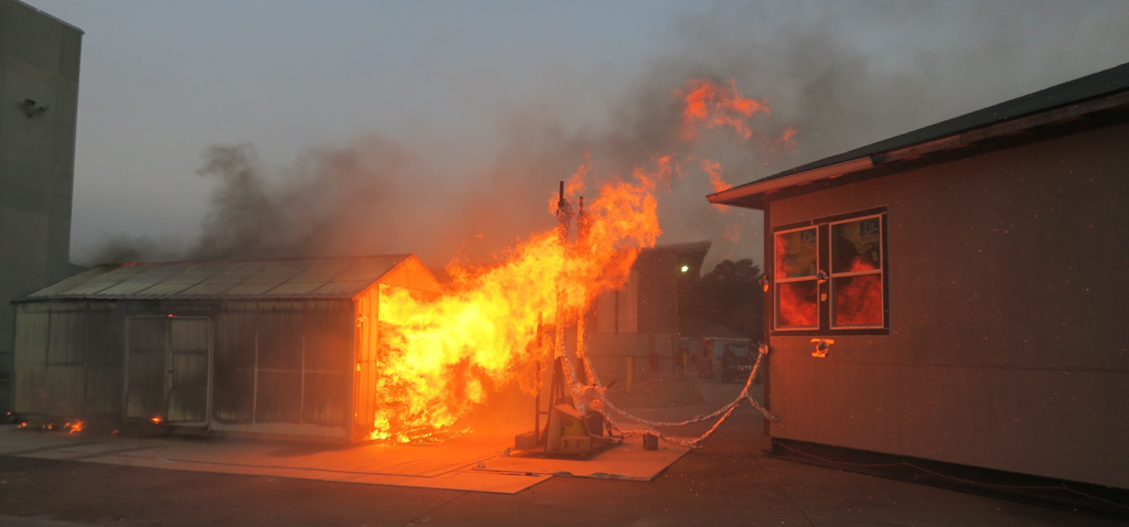 Experiment setup is shown with a test structure, reflective of a California Building Code Chapter 7A-compliant home, exposed to fire from a nearby structure (shed).